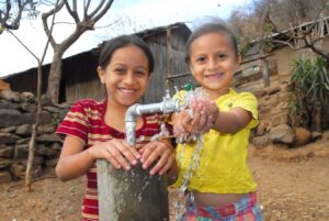 Two children stand next to an outdoor water faucet in Nicaragua. One rests her had on the pump. The other, in a yellow shirt, has her hands in running water.