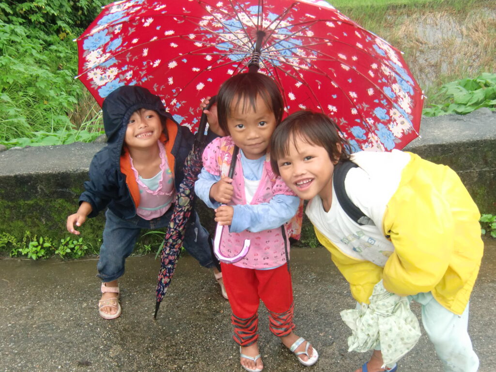 Three small children smile standing under a bright red umbrella