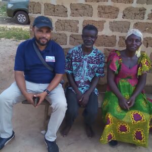 A group of three people sitting on a bench against a brick wall. The person on the left wears a cap and blue shirt, the middle person wears a patterned shirt, and the person on the right wears a colorful dress, all smiling warmly.Project Officer with Samuel and Angelina.jpg